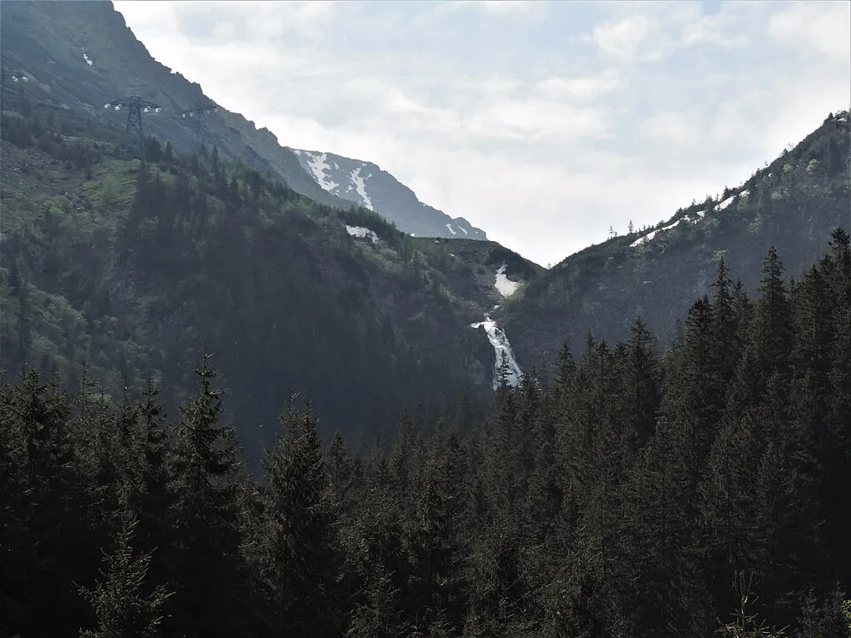Bâlea Waterfall seen from the forested valley below as a thin white cascade dropping down a rocky gully in the Făgăraș Mountains, with patches of late snow on the ridge above.